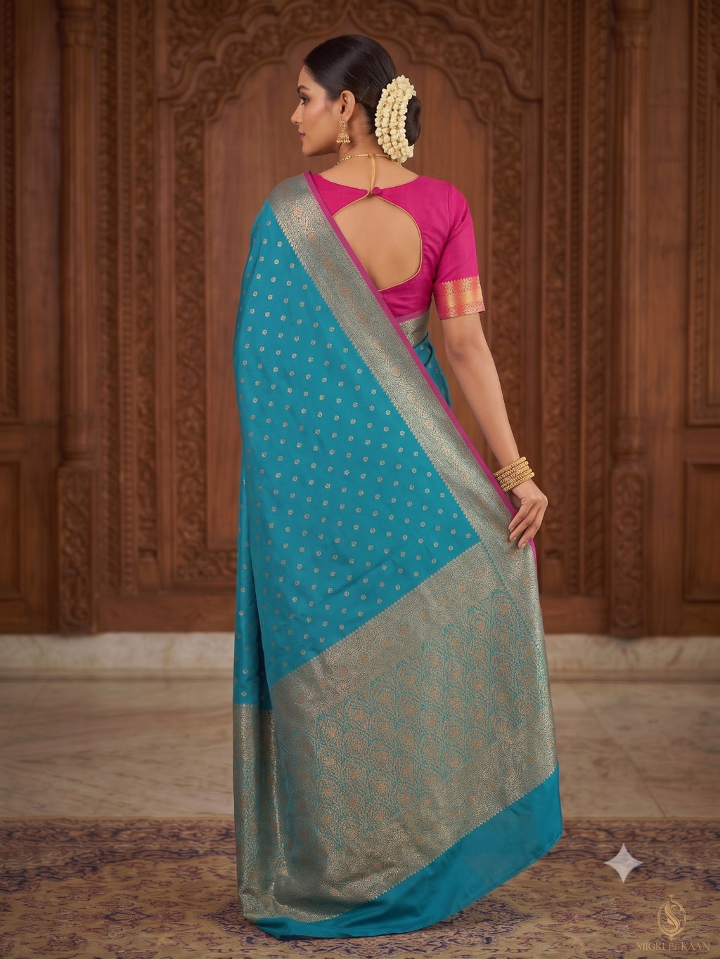 A woman wearing a teal blue and silver saree with a pink blouse, standing in front of a carved wooden wall. The saree has small gold motifs, and the blouse has gold and orange border details.